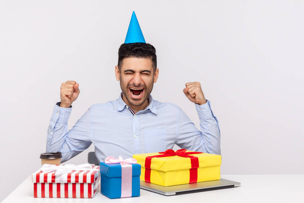 Excited man office employee with party cone sitting workplace surrounded by gift boxes and shouting for joy, celebrating birthday, rejoicing many presents. studio shot isolated on white background - 写真・画像