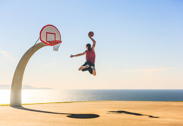 Koszykarz skaczący w powietrzu zdobywając niesamowity energiczny slam dunk na boisku do piłki nożnej ulicy z pięknym widokiem na ocean w tle. Sport, motywacja i elewacja. - Zdjęcie, obraz