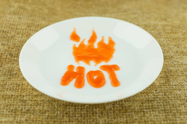 vaarallisen mausteinen chilikastike: sana "kuuma" kirjoitettu valkoiselle lautaselle chilikastikkeella, kirjainten liekit tulevat esiin, näkymä edestä, tavallinen taustakangas, lyhyt tarkennus - Valokuva, kuva