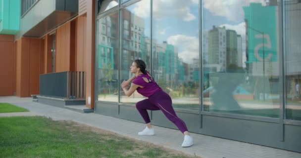 Mujer atlética hace ejercicios de fitness en la hierba cerca de la fachada del edificio de vidrio del complejo de apartamentos, fitness en el entorno urbano, clases de yoga al aire libre, entrenamiento físico en la ciudad, 4k 120p Prores - Metraje, vídeo