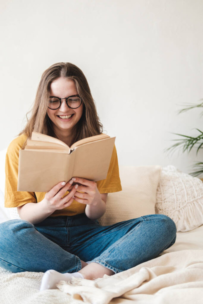 Young beautiful girl student in glasses, an orange T-shirt and blue jeans sits on bed with pillows and reads paper book. Self-education concept at home during quarantine. Home schooling, hobby. - 写真・画像