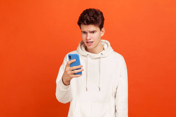 Wondered teenager in white casual sweatshirt with hood looking at smartphone screen with amazed face, bad connection. Indoor studio shot isolated on orange background - Foto, Imagen