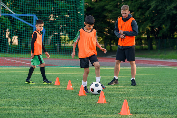 Adolescente persistente en ropa deportiva entrena el fútbol en el campo de fútbol y aprende a rodear la pelota entre los conos de entrenamiento. - Foto, imagen