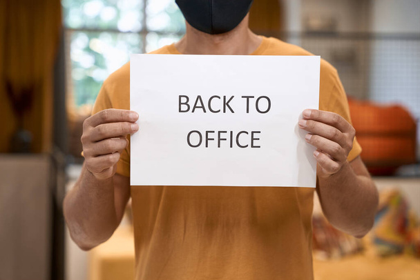 Schnappschuss eines männlichen Büroangestellten mit schwarzer Schutzmaske, der Papier mit Text vor laufender Kamera zeigt - Foto, Bild