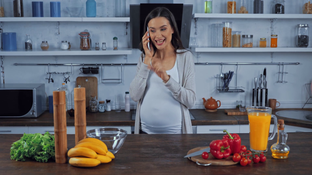 Pensive kobieta w ciąży rozmawia na smartfonie w pobliżu warzyw w kuchni - Materiał filmowy, wideo