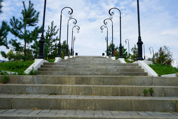 stone steps of a large staircase in a park with beautiful street lamps, a neatly trimmed green lawn and young conifers against a background of blue sky and white clouds on a clear warm summer day - Foto, Imagem
