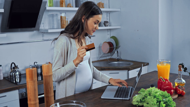 Mujer embarazada usando tarjeta de crédito y portátil cerca de verduras en la mesa en la cocina  - Metraje, vídeo