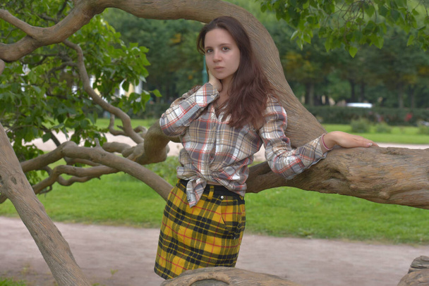 beautiful slender brunette girl in a plaid skirt and blouse in the summer by a tree in the park - Valokuva, kuva