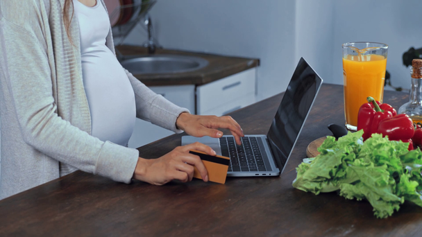 Vista recortada de la mujer embarazada usando el ordenador portátil y la tarjeta de crédito cerca de verduras  - Imágenes, Vídeo