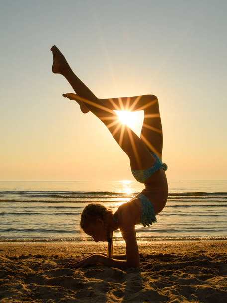 Güneşin ışınları sahildeki bir jimnastikçinin bacaklarında parlıyor. Kız amuda kalkar ve bacaklarını üçgen şeklinde tutar. Denizde gün batımı. - Fotoğraf, Görsel