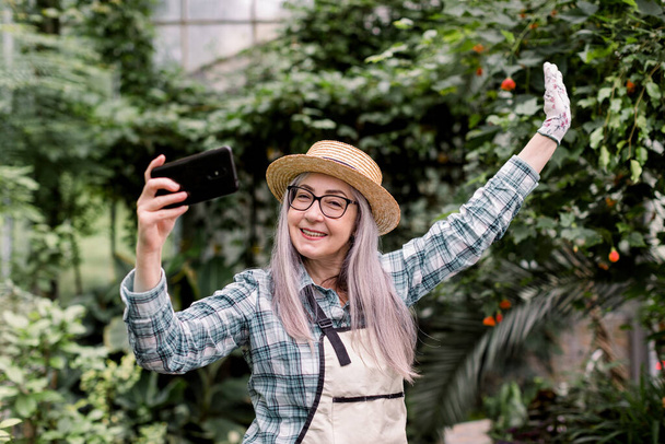 Pleasant positive joyful elderly woman gardener, posing in beautiful greenhouse with exotic plants and palm trees while making selfie photo or video vlog for her followers using her smartphone - Foto, imagen