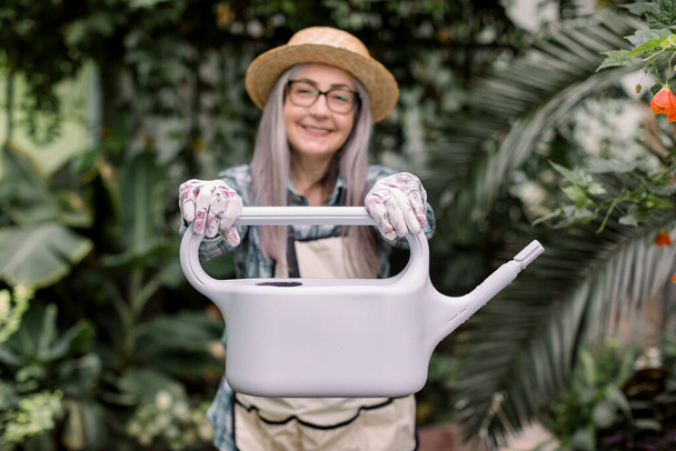 Gardening and greenhouse concept. Beautiful cheerful gray haired senior woman in straw hat and glasses, posing with gray water can for watering plants in floral greenhouse. Focus on the can - Photo, Image