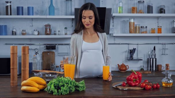 Беременная женщина пьет апельсиновый сок возле овощей на столе  - Кадры, видео