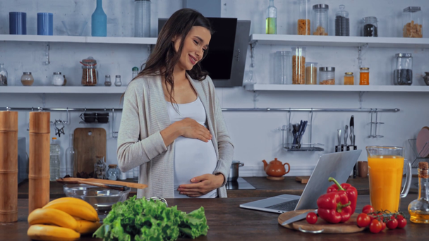 Kobieta w ciąży mając wideo rozmowy na laptopie w pobliżu warzyw na stole w kuchni  - Materiał filmowy, wideo