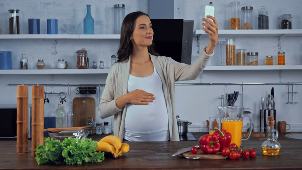 Mujer embarazada tomando selfie con teléfono inteligente cerca de verduras en la cocina  - Imágenes, Vídeo