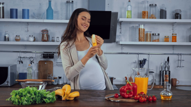 Беременная женщина ест банан возле овощей на кухонном столе  - Кадры, видео