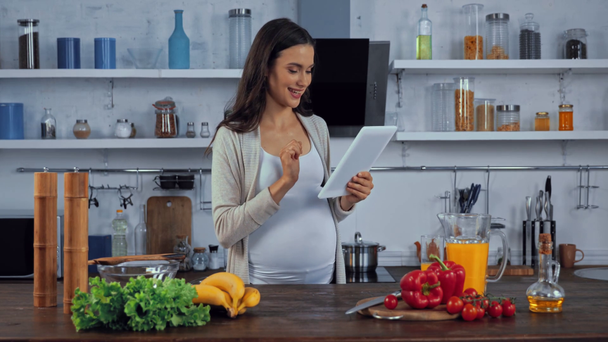 Zwangere vrouw met behulp van digitale tablet in de buurt van verse groenten op keuken tafel  - Video