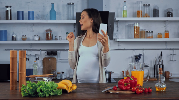 Zwangere vrouw met video bellen op smartphone in de buurt van groenten in de keuken - Video