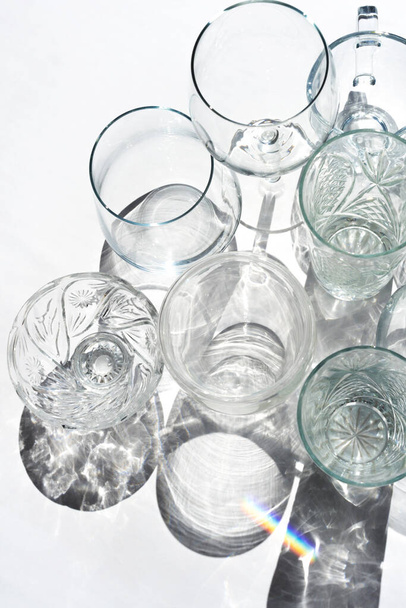 Many empty crystal wine, coctail glasses on white. Transparent beverage cups silhouette on sunlight, rainbow reflection - Photo, Image