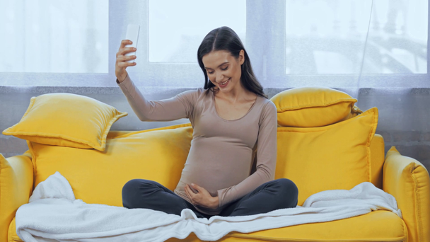 Mujer embarazada tomando selfie con smartphone en la sala de estar  - Metraje, vídeo