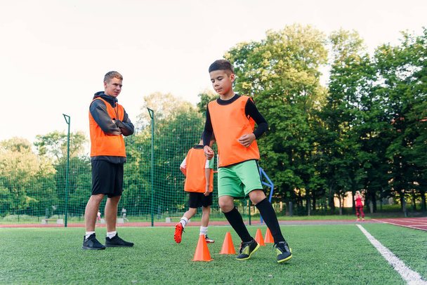 Motivado deportivo 13-s chicos en chalecos naranjas corriendo entre los conos de plástico durante el entrenamiento de fútbol en el estadio. - Foto, Imagen