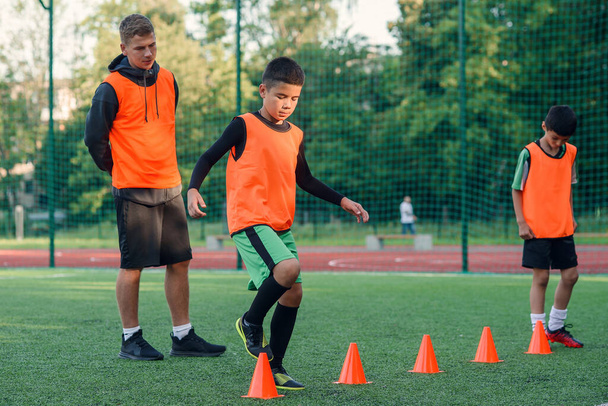 Motivated sporty 13-s boys in orange vests running among the plastic cones during soccer training at the stadium. - Fotografie, Obrázek