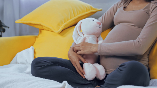 Vista ritagliata della donna incinta che tiene il giocattolo morbido e tocca la pancia a casa - Filmati, video