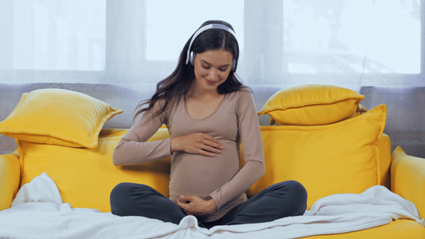 Pregnant woman listening music in headphones on couch  - Footage, Video