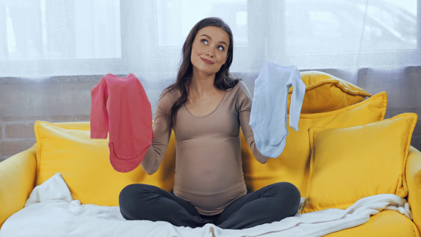 Έγκυος γυναίκα που κρατάει μωρουδιακά ρούχα στον καναπέ  - Πλάνα, βίντεο