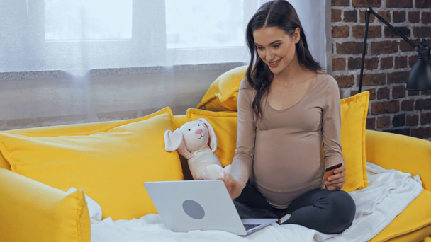 Mujer embarazada usando portátil y tarjeta de crédito en el sofá  - Imágenes, Vídeo