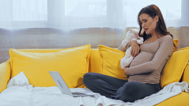 Pregnant woman with soft toy crying near laptop on couch  - Felvétel, videó
