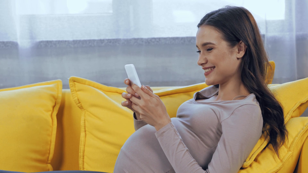 Έγκυος γυναίκα που χρησιμοποιεί το smartphone στον καναπέ στο σπίτι  - Πλάνα, βίντεο