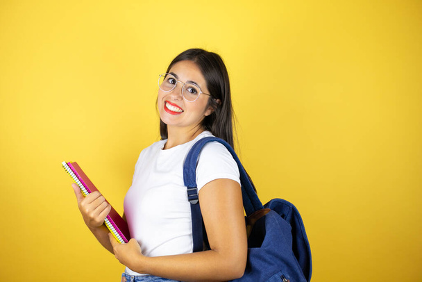 Молода красива студентка в рюкзаку тримає блокнот над ізольованим жовтим тлом, посміхаючись і дивлячись на камеру
 - Фото, зображення