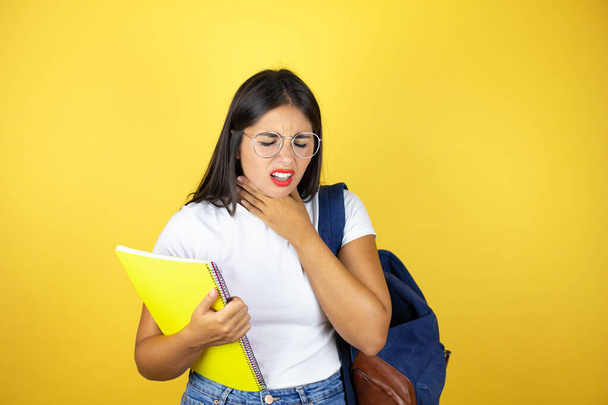 Młoda piękna studentka nosząca plecak trzymająca notatnik na odizolowanym żółtym tle dotykająca bolesnej szyi, bólu gardła na grypę, chrząszcz i infekcję - Zdjęcie, obraz