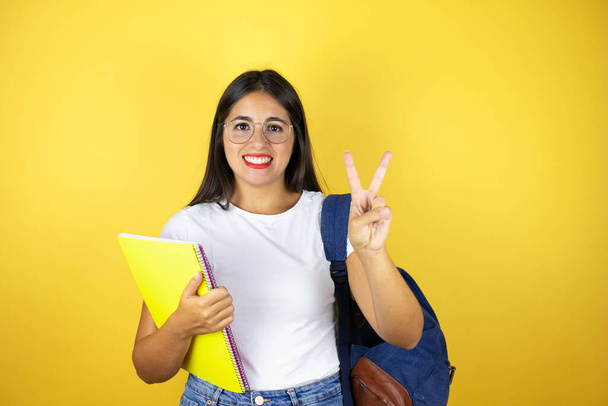 Νεαρή όμορφη φοιτήτρια γυναίκα φορώντας σακίδιο κρατώντας σημειωματάριο πάνω από απομονωμένο κίτρινο φόντο δείχνει και δείχνει προς τα πάνω με τα δάχτυλα νούμερο δύο, ενώ χαμογελά αυτοπεποίθηση και χαρούμενος - Φωτογραφία, εικόνα