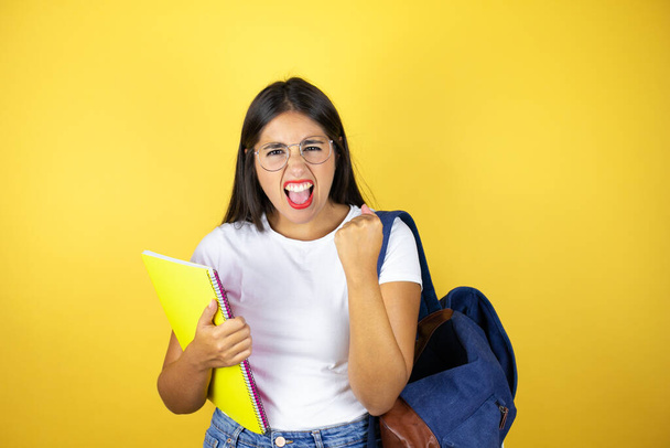 Jovem mulher estudante bonita usando mochila segurando notebook sobre fundo amarelo isolado muito feliz e animado fazendo gesto vencedor com braços levantados, sorrindo e gritando para o sucesso. - Foto, Imagem