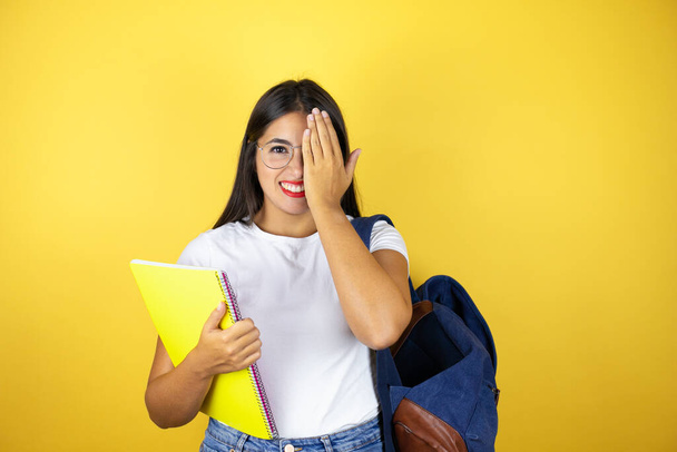 Молода красива студентка в рюкзаку тримає блокнот над ізольованим жовтим тлом, що покриває одне око рукою, впевнена посмішка на обличчі та несподівані емоції
. - Фото, зображення