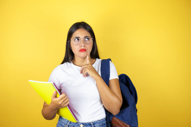 Junge schöne Studentin mit Rucksack, Notizbuch über isoliertem gelben Hintergrund denkend und zur Seite schauend - Foto, Bild
