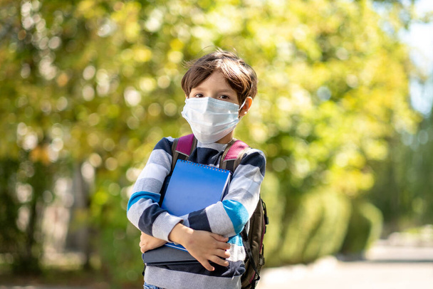 Coronavirus COVID-19 quarantine pandemic, portrait of a schoolboy wearing a protective mask outdoors in the open air. Child Little boy with a backpack going to school. Education. Health concept. Slow motion - 写真・画像