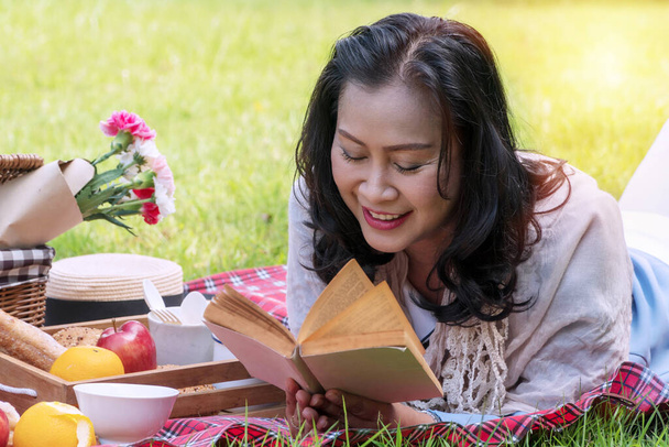 Elderly lifestyle concept. Senior women 50s reading attractive book in her hand have leisure time with picnic have apple, oranges, milk and bread on green grass in the public park . - Photo, Image