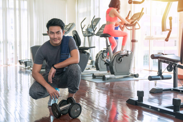 Muskularny młody mężczyzna z hantlem i ręcznikiem odpoczywający po treningu z rozmytą młodą kobietą szczupłą sylwetką na tle urządzeń fitness w siłowni. Koncepcja ćwiczeń i sportu - Zdjęcie, obraz
