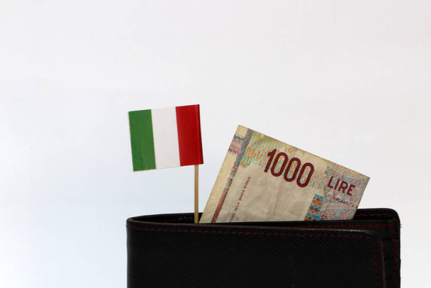 Χαρτονόμισμα χιλίων λιρετών Ιταλίας και σημαία της μίνι Ιταλίας κολλάνε στο μαύρο πορτοφόλι με λευκό φόντο. Έννοια της χρηματοδότησης ή του νομίσματος. - Φωτογραφία, εικόνα