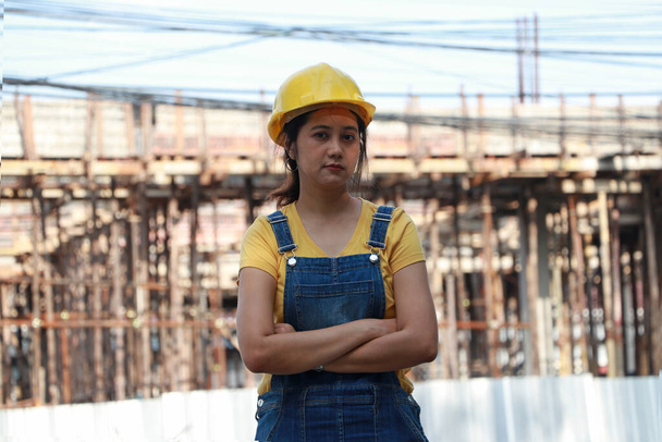 Engenheira civil ou arquiteta com capacete amarelo, em pé e abraçando peito na área de trabalho. - Foto, Imagem