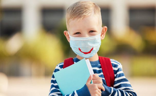 Optimistic smart first grade schoolboy in casual wear and eyeglasses wearing protective mask with drawn smile holding copybooks and looking at camera against blurred urban backgroun - Photo, Image
