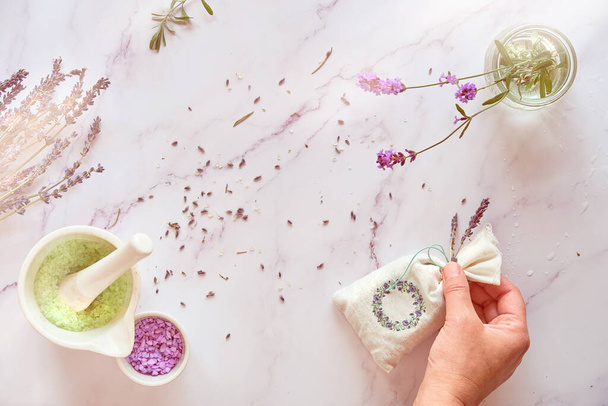 Handmade lavender sachets and home made sugar scrub. Hand holds linen bag with dry lavender flowers. Top view on white marble table. - Foto, Imagem