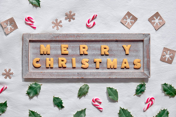 ext Merry Christmas made with cookies on rustic wooden board. Geometric flat lay on white textile background. Eco-friendly decorations from craft paper , candy canes and natural holly leaves. - Фото, зображення