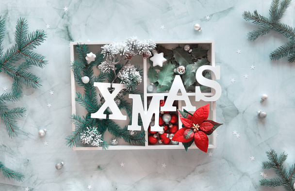 Xmas decorations in white box. Christmas flat lay on marble table. Natural fir twigs, holly leaves and glass trinkets. Letters Xmas on top of the show box. Wintertime background in green, white, red - Fotó, kép