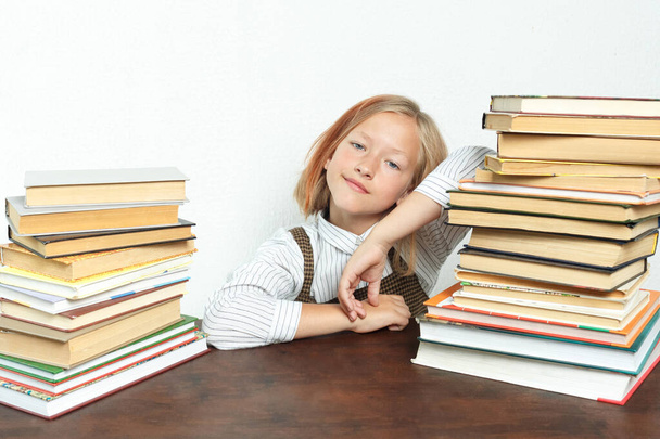 Portrait of a teenage girl who sits at a table among the books. Looks into the camera. Isolated background. Education concept. - Foto, Imagem