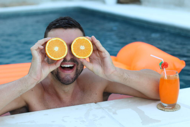 közeli kép egy jóképű fiatalemberről, aki fél narancsot tart a szeme előtt a medencében. - Fotó, kép