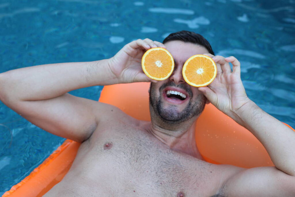 close-up retrato de belo jovem flutuando no colchão inflável segurando metades de laranja na frente dos olhos na piscina - Foto, Imagem